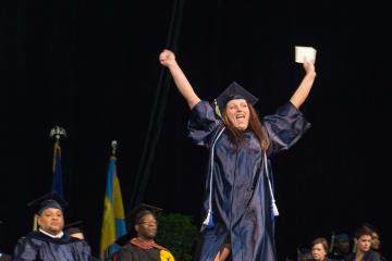A graduate walks across the stage and raises her arms in celebration.