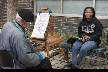 A student smiles as she sits and has her portrait drawn.